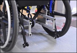 Rear view of manual wheelchair showing UDIG attachments. Two vertical posts are located between the rear wheels, and attached to the frame with horizontal rods at the top and bottom that extend back to near the tiedown hooks.