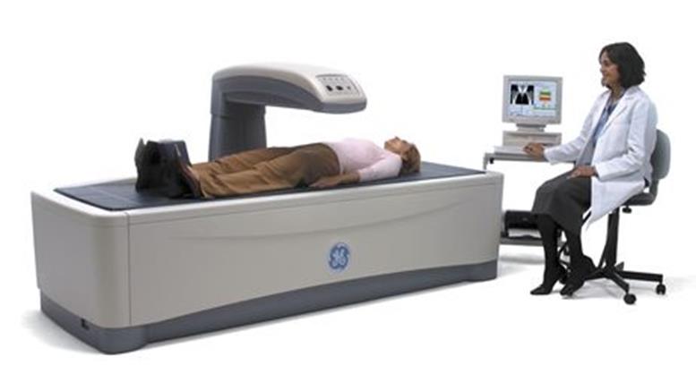 Picture shows a Dual Energy X-ray Absorptiometry (DXA) system for Osteoporosis assessment with a patient lying on the scanning bed.  A technician is seated beside the equipment viewing a computer monitor. 