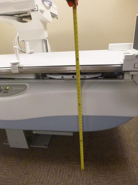 Picture of an Angulating Radiographic and Fluoroscopic Exam Table whose fixed height is approximately 34.5 inches.  Tape measure shown beside transfer surface to show height.  Also note the equipment imaging components on the opposite side of where the patient transfers.