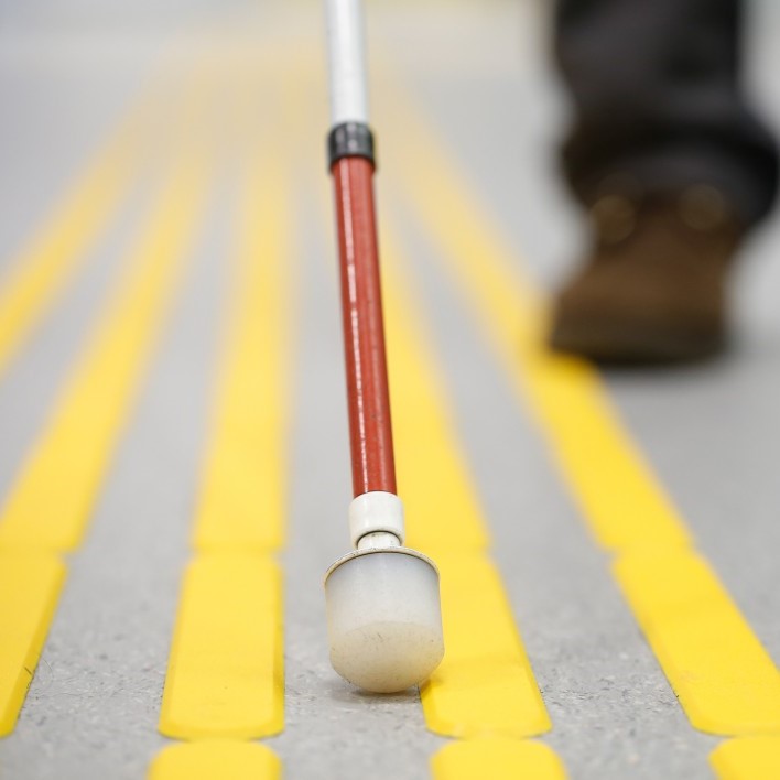cane trailing sidewalk (photo closeup on cane with red stripe and white bulb at end, in front of a blurred shoe)