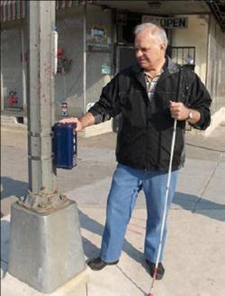A man stands on a corner beside a pole, with a white cane in his left hand and his right hand on top of an APS on the pole.