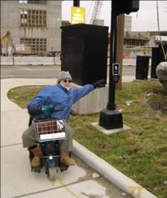 At an intersection corner, a man on a scooter is leaning far to his left to try to reach a pushbutton that is located in a grassy area behind a raised curb. It is a couple of feet out of reach.