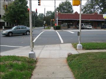 Photo of crossing, looking across street from sidewalk, with street parallel to photographer on left. Landscape strips are located between the sidewalks and streets. Pushbutton with arrow pointing toward the street in front of the photographer is on a pole on the right side of the photo, in the landscape strip at the edge of the sidewalk and landing for the street in front of the photographer. Another pedestrian signal pole is located on the left side of the photo, in the landscape strip beside the curb ramp for the street to the left in the photo. Sign and pushbutton are on the back side of pole, barely visible in photo.
