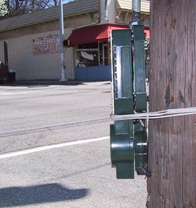 APS that was damaged in a crash and temporarily reinstalled with wire. Description:  Photo with pole on right and intersection visible to left.  Several strands of wire are wrapped around the pole and APS unit.  The APS aims diagonally across intersection.