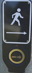 Photo of black rectangular unit with a sign with a print arrow and pedestrian signal. On the bottom part of the unit is a large pushbutton with an arrow.