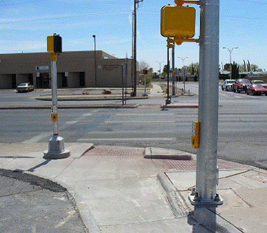 Photo of crossing, looking across street from sidewalk, with street parallel to photographer on right.  Pushbutton with arrow pointing toward the right side of the photo (toward parallel street) is on a pole on the left side of the photo, beside the landing and curb ramp for the street in front of the photographer.  Another pushbutton is located on a large signal pole in forefront of the photo, with arrow pointing toward street in front of photographer, close to the ramp for the parallel street, but more than 15 feet from the street the arrow points toward.