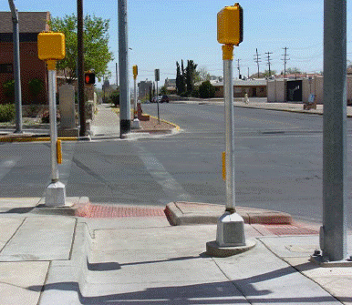 Photo of crossing, looking across street from sidewalk, with street parallel to photographer on right. Pushbutton with arrow pointing toward the street in front of the photographer is on a pole on the left side of the photo, beside the landing and curb ramp for the street in front of the photographer. Another pedestrian signal pole is located on the right side of the photo, beside the curb ramp for the street to the right in the photo.