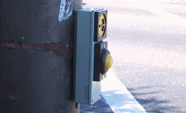 On left side of photo, APS pushbutton unit with tactile arrow is mounted on a large pole. Crosswalk line extends across street slightly to left; arrow angles right toward center of intersection.