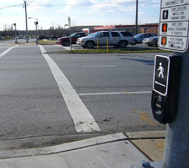 On right side of photo, APS pushbutton unit with tactile arrow is mounted on a large pole. Crosswalk line extends across street in line with arrow and pushbutton unit.