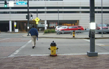 Photo from sidewalk looking across street with street parallel to photographer on left. Pedestrian is crossing the street. Pole on right in photo, with two pushbuttons on it, is over 10 feet to the right of the crosswalk line. A fire hydrant is located between pole and crosswalk.