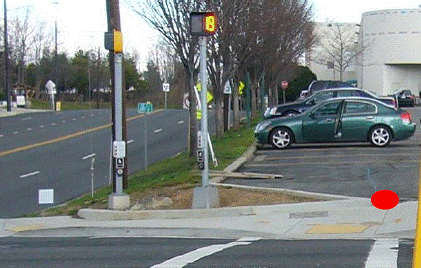 View of corner from the middle of the crosswalk, approaching the corner. Curb ramp is directly in the middle of the crosswalk with a steep cross slope from the right; both pedestrian signal poles and pushbuttons are to the left of the crosswalk behind raised curbing at the back of the sidewalk. An APS for the crosswalk is visible with arrow pointing toward the right of the photo (should be aligned toward the photographer).