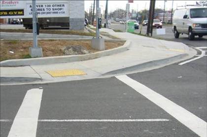 View of corner from the middle of the crosswalk, looking West, with parallel street to photographer's right. APS are visible on pole on left side of crosswalk behind the sidewalk, and on another pole to the right of the crosswalk, also behind the sidewalk. Arrow on APS on right aims northwest. Sidewalk slopes down to a perpendicular ramp with a good deal of cross slope.