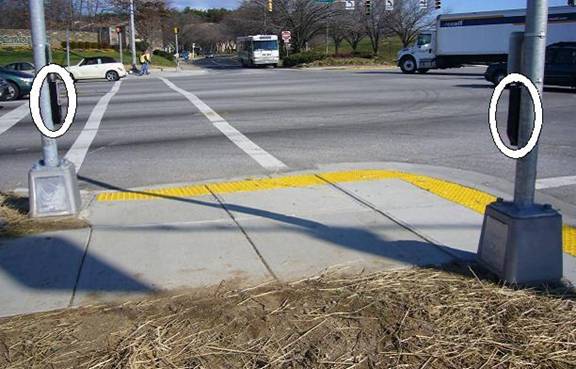 Photo of corner, looking across a street, with parallel street on photographer's right. APS for crosswalk straight ahead is on a pole to the left of the curb ramp, in line with the crosswalk line. APS for crosswalk to right is on pole on right side of photo, next to the landing for the curb ramp for that crossing.