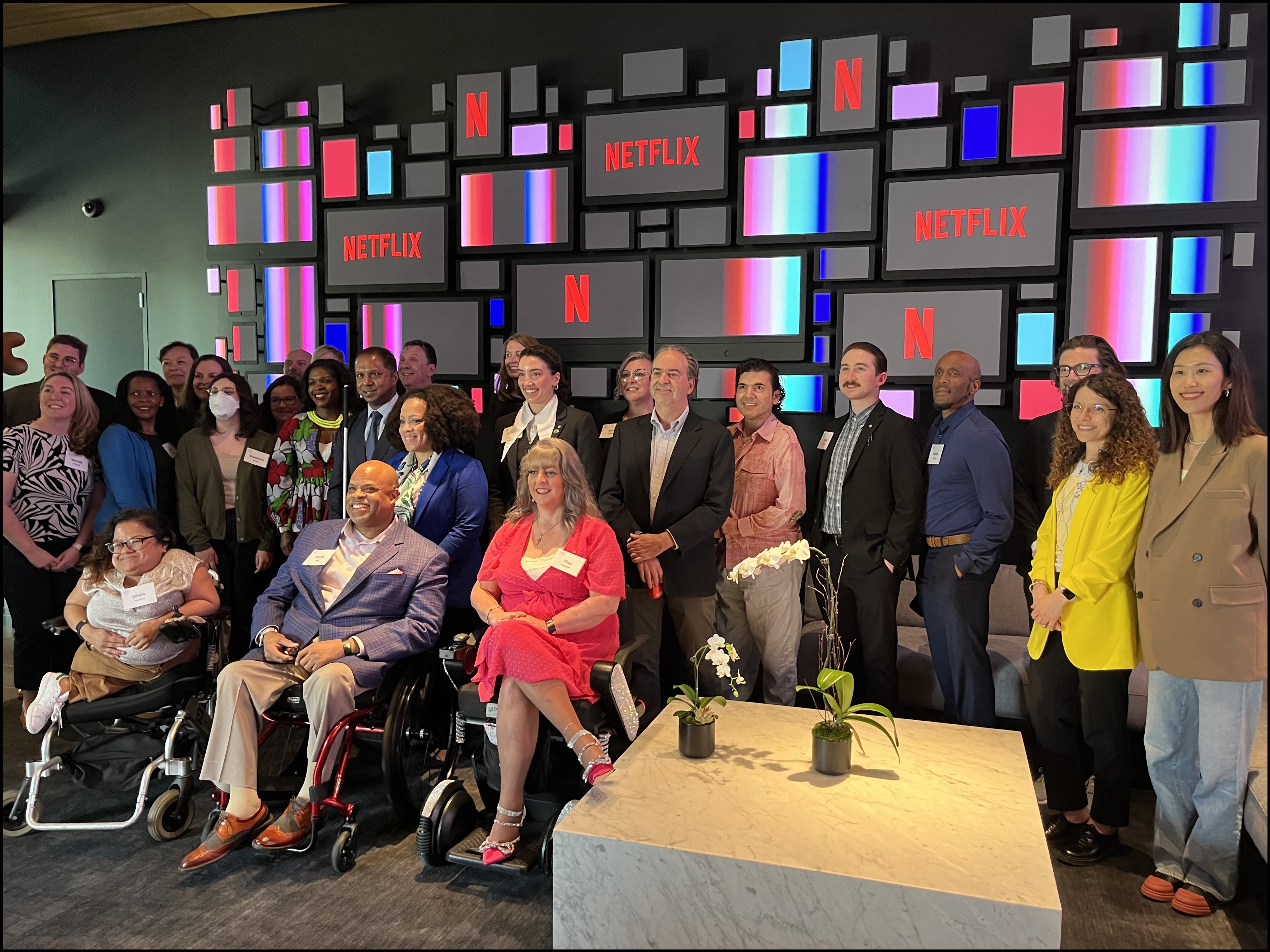 A large group of Access Board Members and staff, and Netflix staff members pose in the lobby of Netflix's Los Angeles office. Behind them is a wall of screens with variations of the Netflix logo appearing throughout.