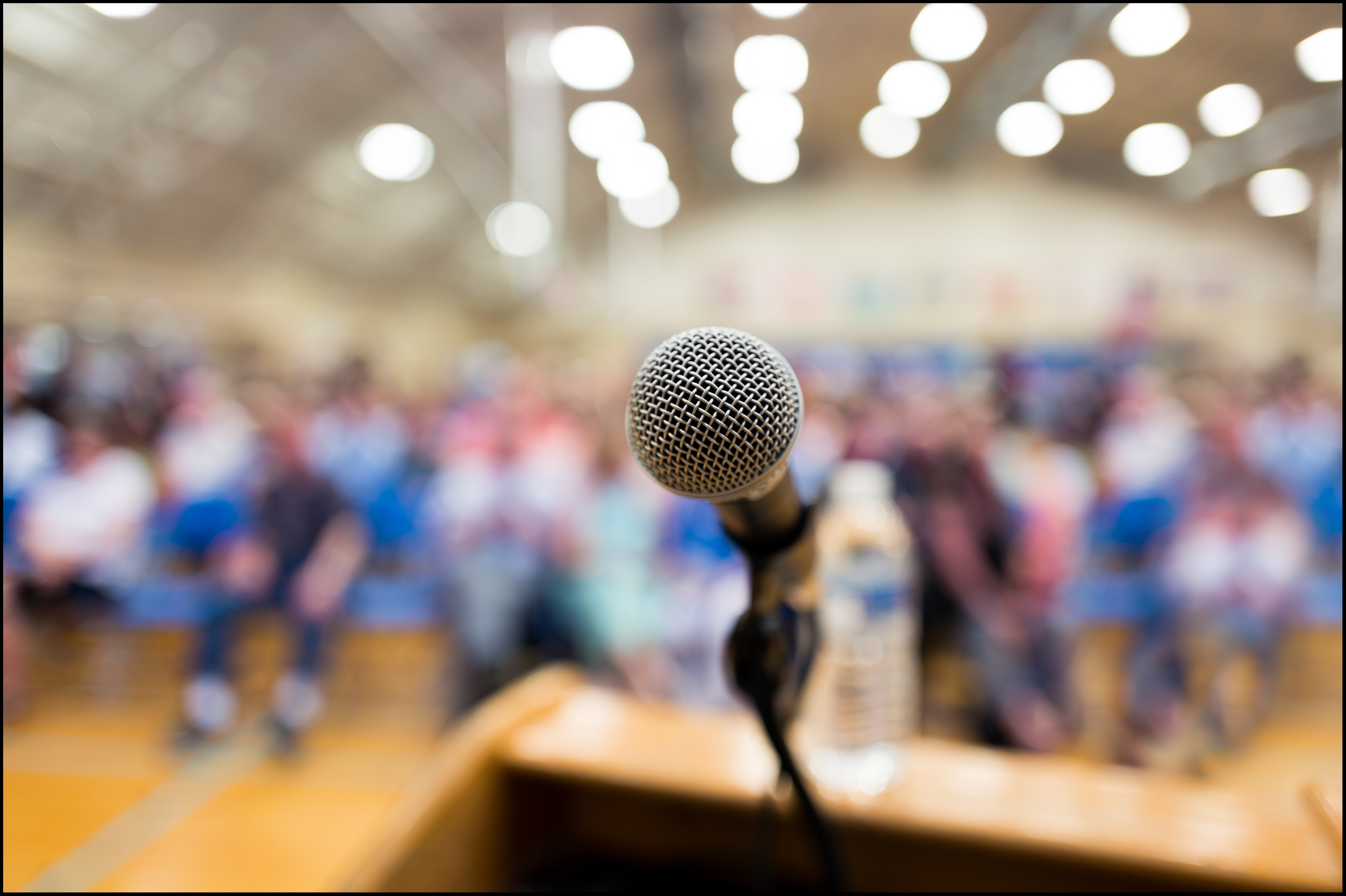 A microphone connected to a podium stands before a seated audience. The microphone is in focus, and the audience is out of focus. The floor of the room is a light colored wood, and the ceiling is adorned with numerous rows of lights.