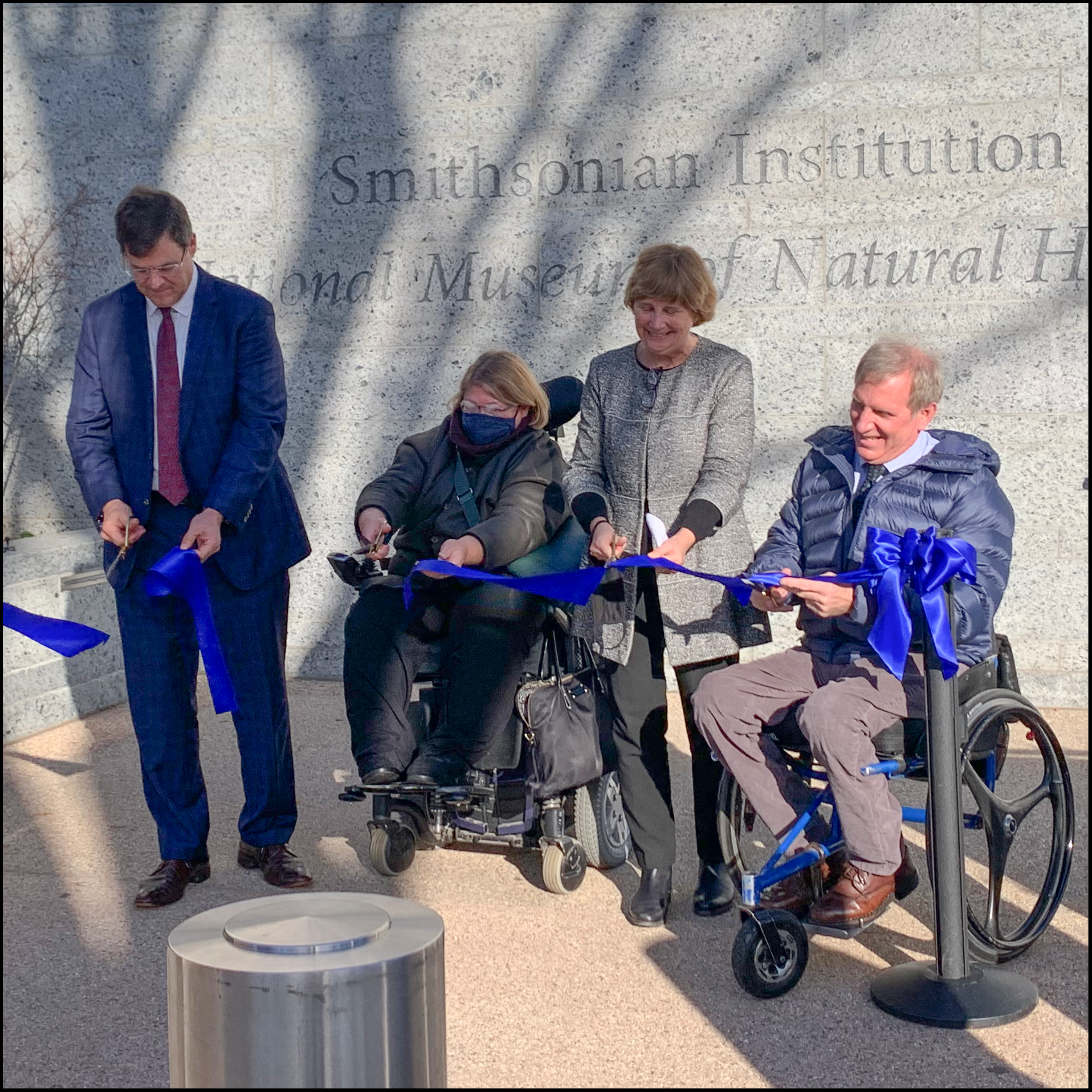 Photo of four people outside holding blue ribbon and scissors, Smithsonian Instituion is engraved in stone behind them.
