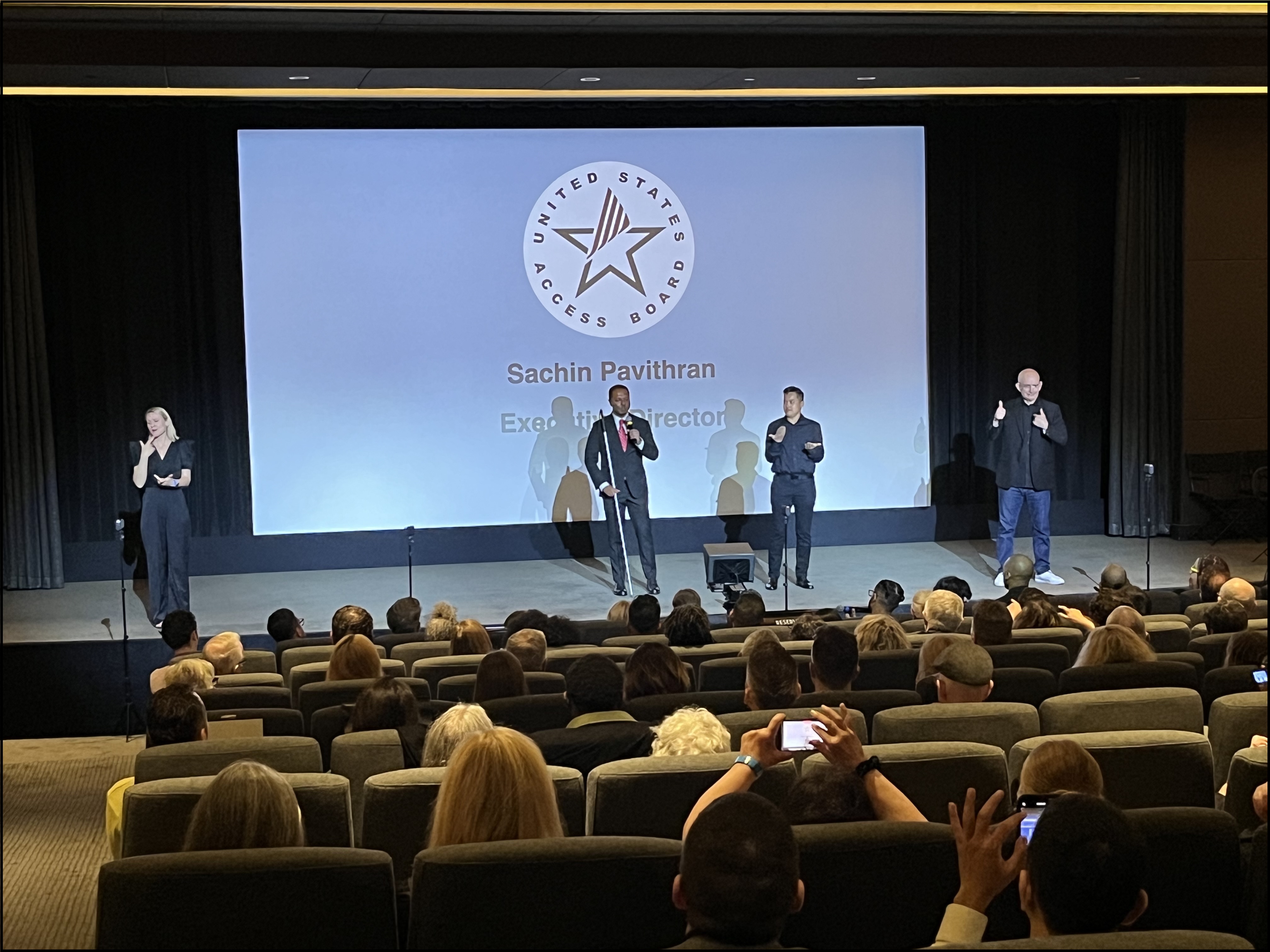 Access Board Executive Director Sachin Pavithran stands on stage at the opening ceremony of the SignLight International Film Festival. On either side of the stage are sign language interpreters, and on a screen behind him is the Access Board logo and his title.
