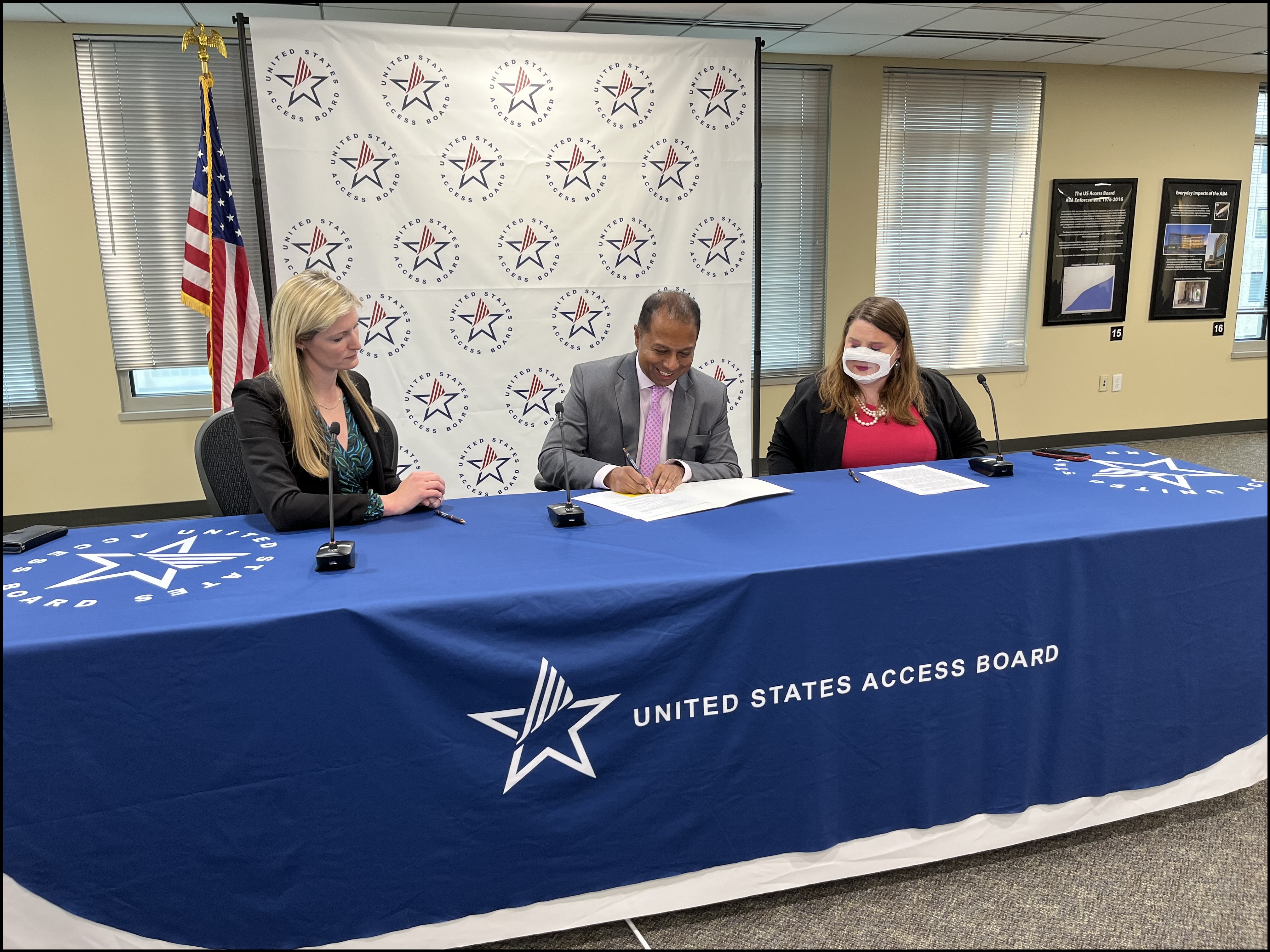 Access Board Executive Director Sachin Pavithran, President and CEO of AAPD Maria Town, and President and CEO of CDT Alexandra Reeve Givens sign the MOU. They sit at a table with a blue tablecloth that has the Access Board logo on it. Behind them is a banner with a repeating pattern of the Board's logo next to the flag of the United States.